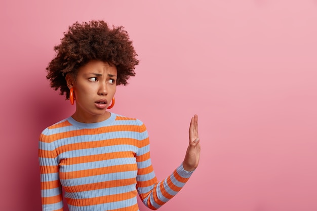 Photo gratuite le mécontentement femme afro-américaine garde la paume dans le geste de refus, dit reste loin de moi et ne se rapproche pas, évite les endroits bondés, grimaces déçues, isolé sur un mur rose, espace vide