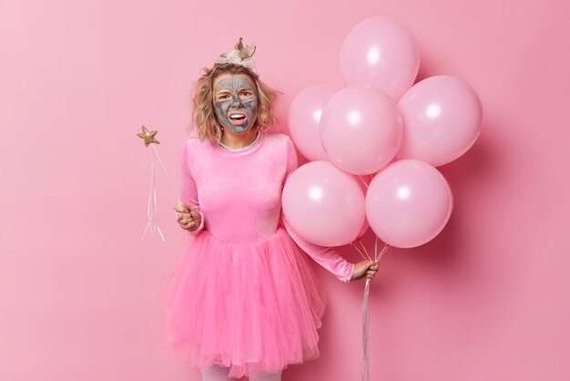 Mécontentement indigné, la jeune femme a l'air perplexe applique un masque d'argile nourrissant sur le visage entend des nouvelles désagréables se prépare pour la fête et la célébration isolée sur fond rose tient des ballons gonflés