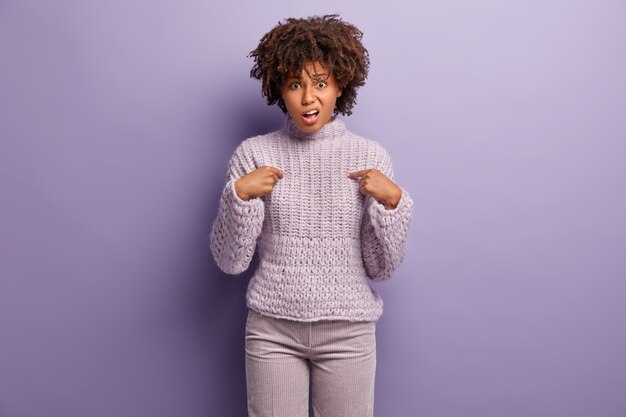 Le mécontentement indigné de la jeune femme se pointe du doigt, demande pourquoi c'est son devoir, fronce les sourcils avec mécontentement, exprime son désaccord, porte un pull en tricot, isolé sur un mur violet. Pourquoi moi?