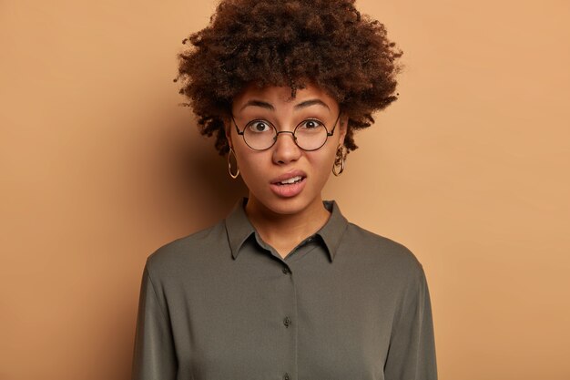 Mécontentement, jeune femme afro-américaine exprime le mépris et l'incrédulité, sourit avec mécontentement, porte des lunettes optiques transparentes et une chemise, pose à l'intérieur contre un mur marron