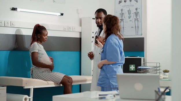 Médecin afro-américain donnant des pilules sur ordonnance à une patiente enceinte lors d'une visite de contrôle. Médecin de sexe masculin aidant une femme avec un ventre de grossesse, montrant une bouteille de traitement médicamenteux.