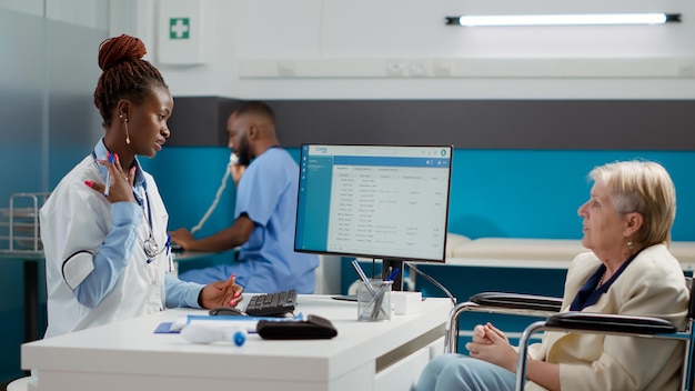 Photo gratuite médecin afro-américain faisant une visite de contrôle avec une utilisatrice de fauteuil roulant, parlant de handicap physique et de diagnostic de soins de santé. consultation d'une femme handicapée lors d'un rendez-vous médical.