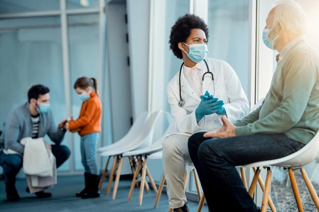 Médecin afro-américain et homme âgé communiquant dans la salle d'attente de l'hôpital tout en portant des masques faciaux