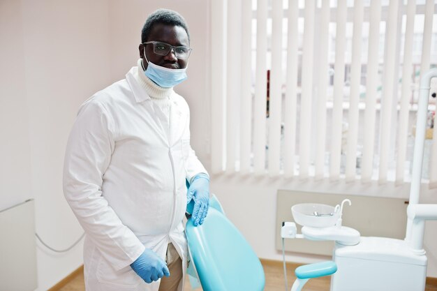 Médecin afro-américain portant des lunettes et un masque debout près de la chaise du dentiste dans une clinique dentaire