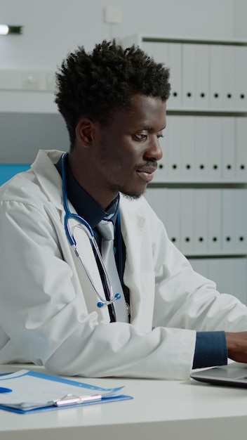 Photo gratuite médecin afro-américain utilisant un ordinateur portable assis au bureau dans la chambre d'hôpital. spécialiste de la santé noir travaillant avec la technologie pour la consultation dans une armoire moderne dans un établissement médical