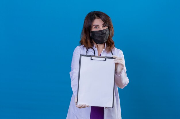 Médecin d'âge moyen portant une blouse blanche dans un masque facial de protection noir et avec un stéthoscope montrant le presse-papiers