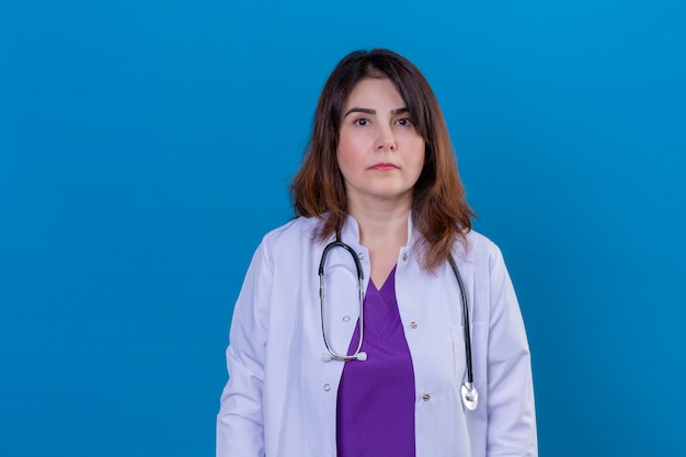 Médecin d'âge moyen portant un manteau blanc et avec un stéthoscope avec une expression de confiance grave sur mur bleu isolé