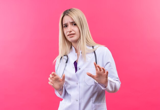 Médecin confus jeune fille blonde portant un stéthoscope en robe médicale a soulevé les mains sur un mur rose isolé