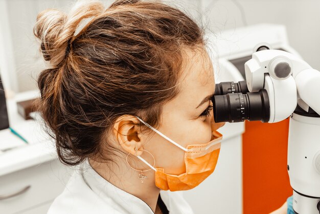 Médecin dentiste de jeune femme regarde à travers un microscope professionnel dans une clinique dentaire. un médecin dans un masque médical et un bonnet jetables. équipement avancé en dentisterie