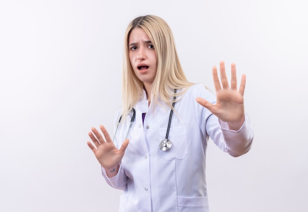 Médecin effrayé jeune fille blonde portant un stéthoscope et une robe médicale montrant le geste d'arrêt avec les deux mains sur fond blanc isolé