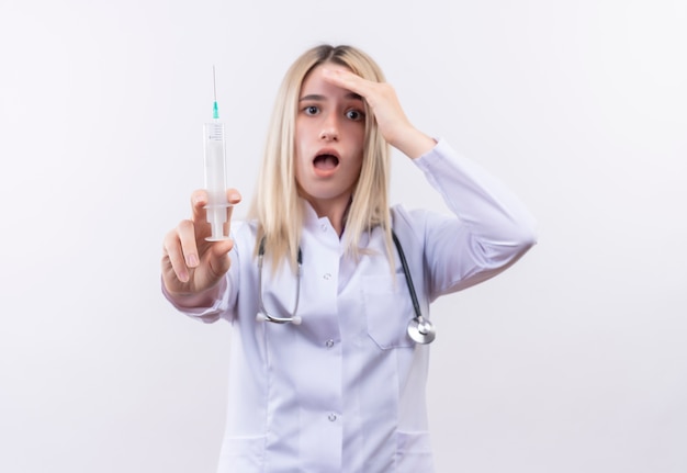 Médecin effrayé jeune fille blonde portant un stéthoscope et une robe médicale tenant une seringue à la caméra a mis sa main sur le front sur fond blanc isolé
