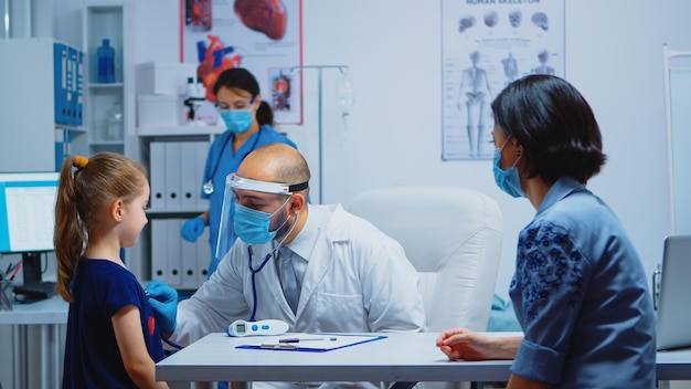 Médecin examinant une fille par stéthoscope dans un cabinet médical pendant la pandémie. Pédiatre spécialiste en médecine avec masque fournissant des services de soins de santé, consultation, traitement en cabinet hospitalier.