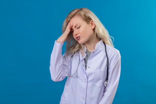 Médecin fatigué jeune fille portant un stéthoscope en robe médicale mis la main sur le front sur fond bleu