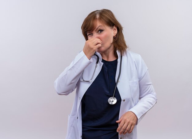 Médecin de femme d'âge moyen ludique portant une robe médicale et un stéthoscope mettant la main sur le nez sur un mur blanc isolé avec copie espace