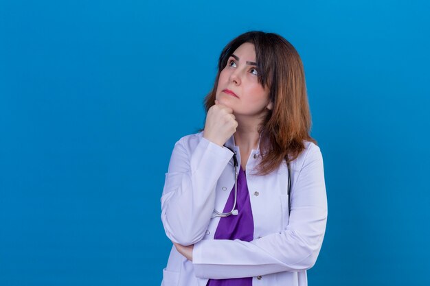 Médecin de femme d'âge moyen portant un manteau blanc et avec stéthoscope debout avec la main sur le menton à la recherche d'expression pensive sur fond bleu