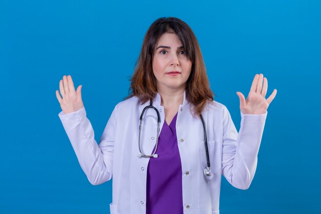 Médecin de femme d'âge moyen portant un manteau blanc et avec stéthoscope en levant les mains dans l'abandon à la confusion debout sur fond bleu