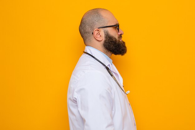 Médecin homme barbu en blouse blanche avec stéthoscope autour du cou portant des lunettes debout sur le côté avec un visage sérieux sur fond orange