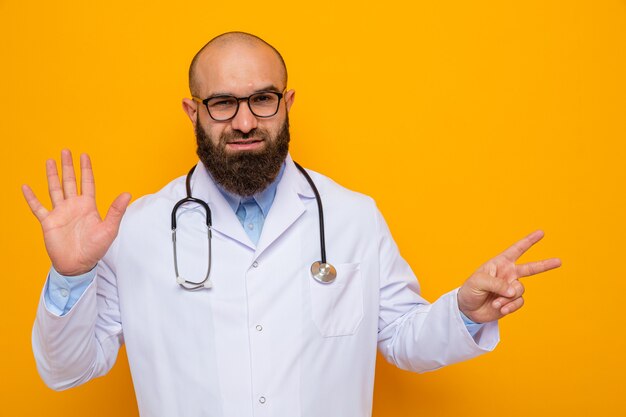Médecin homme barbu en blouse blanche avec stéthoscope autour du cou portant des lunettes heureux et positif montrant cinquième avec la paume pointant avec l'index sur le côté