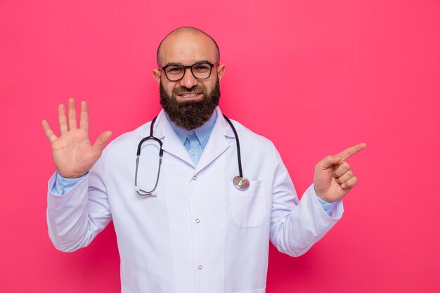 Médecin homme barbu en blouse blanche avec stéthoscope autour du cou portant des lunettes montrant cinquième avec paume pointant avec l'index sur le côté souriant confiant