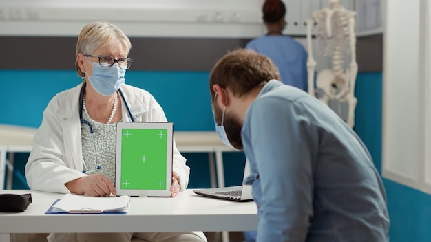 Médecin et homme regardant une tablette numérique avec écran vert dans un cabinet médical. Espace de copie de maquette vierge avec modèle de clé chroma isolé et arrière-plan affiché pendant la pandémie de covid 19.