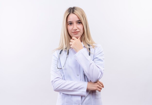 Médecin jeune fille blonde portant un stéthoscope et une robe médicale a mis sa main sur le menton sur un mur blanc isolé