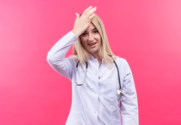 Médecin jeune fille blonde portant un stéthoscope en robe médicale en orthèse dentaire a mis sa main sur le front sur un mur rose isolé