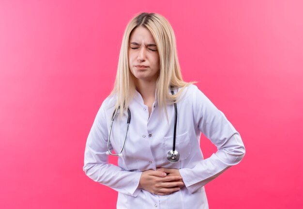 Médecin malade jeune fille blonde portant un stéthoscope en robe médicale a saisi l'estomac douloureux sur fond rose isolé