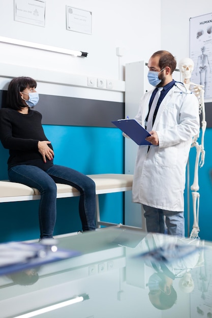 Médecin avec masque facial examinant une patiente enceinte dans un cabinet médical. Femme attendant un enfant et un médecin lors d'une consultation de contrôle pendant la pandémie de coronavirus. Examen de grossesse