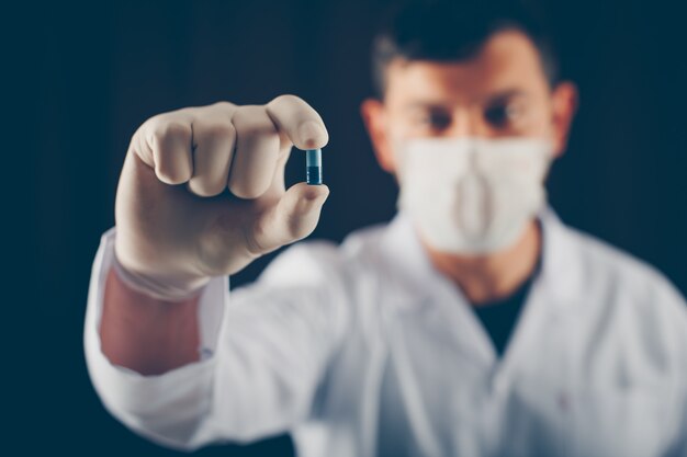 Médecin avec masque et gants tenant une pilule bleue avec ses doigts. vue de côté.