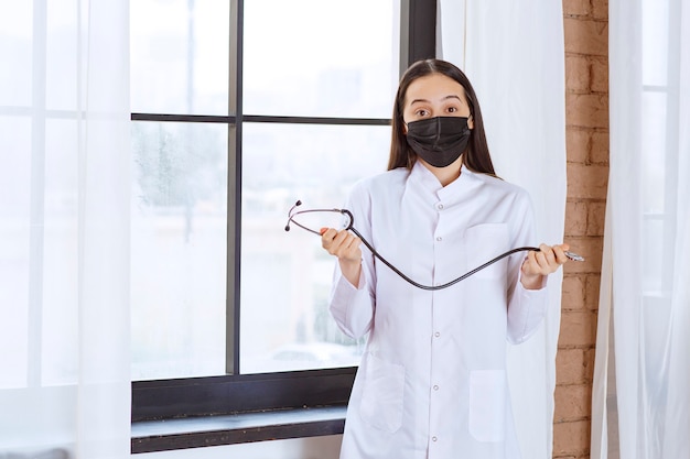 Médecin en masque noir avec un stéthoscope debout près de la fenêtre et semble confus.