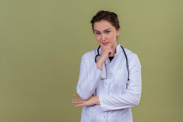 Médecin de la pensée jeune fille portant une blouse médicale portant un stéthoscope a saisi la mâchoire sur fond vert