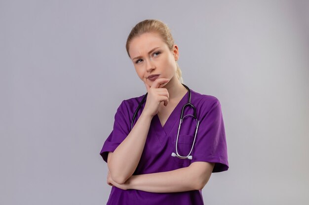 Médecin de la pensée jeune fille portant une robe médicale violette et un stéthoscope a mis sa main sous le menton sur fond blanc isolé