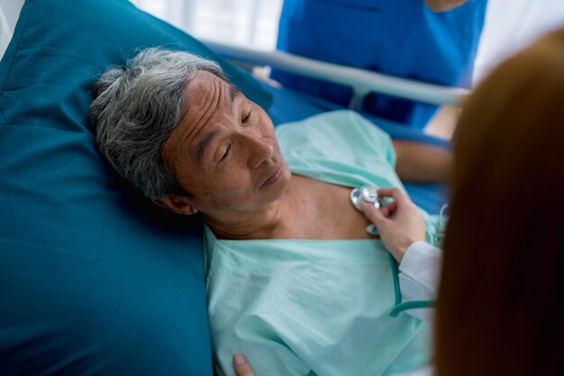 Médecin avec presse-papiers vérifiant et discutant avec un vieil homme asiatique patient allongé sur son lit à l'hôpital avec soin et bonheur