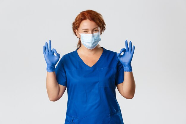 Médecin Rousse Souriant Confiant, Infirmière En Masque Médical, Gants, Montrant Un Geste Correct, Garantir Un Contrôle Sûr Et De Qualité à La Clinique