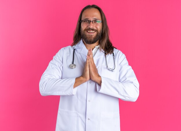 Médecin de sexe masculin adulte joyeux portant une robe médicale et un stéthoscope avec des lunettes regardant la caméra en gardant les mains ensemble isolés sur le mur rose