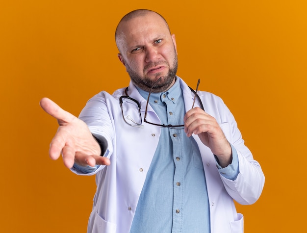Médecin de sexe masculin d'âge moyen agacé portant une robe médicale et un stéthoscope tenant des lunettes tendant la main vers l'avant en regardant l'avant isolé sur un mur orange