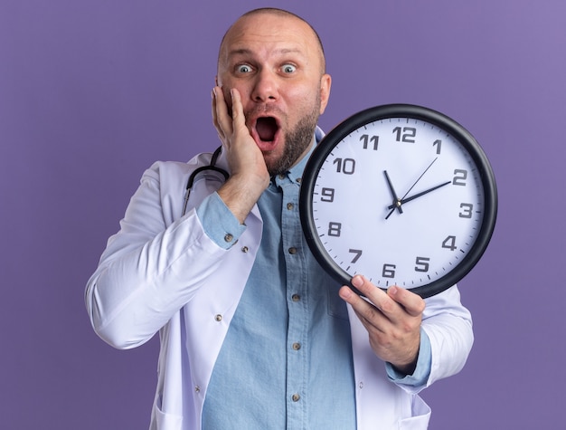 Médecin de sexe masculin d'âge moyen choqué portant une robe médicale et un stéthoscope tenant une horloge en gardant la main sur le visage