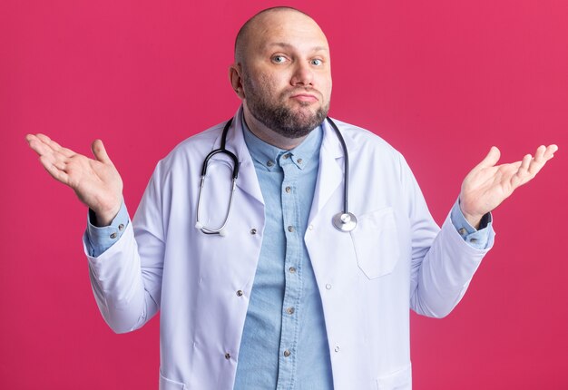 Médecin de sexe masculin d'âge moyen sans idée portant une robe médicale et un stéthoscope faisant je ne sais pas un geste isolé sur un mur rose