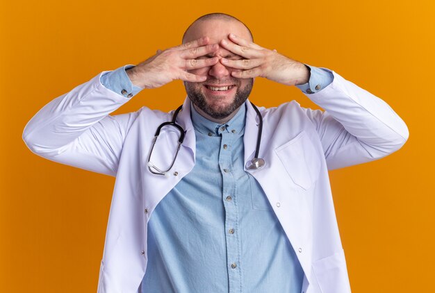 Médecin de sexe masculin d'âge moyen souriant portant une robe médicale et un stéthoscope gardant les mains sur les yeux
