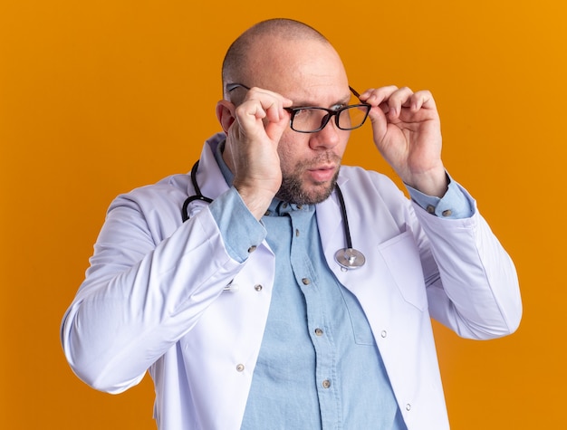 Médecin de sexe masculin d'âge moyen surpris portant une robe médicale et un stéthoscope avec des lunettes saisissant des lunettes regardant le côté isolé sur un mur orange