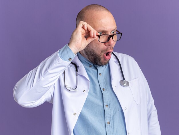 Médecin de sexe masculin d'âge moyen surpris portant une robe médicale et un stéthoscope avec des lunettes saisissant des lunettes regardant le côté isolé sur un mur violet