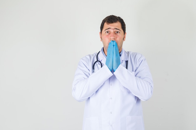 Médecin de sexe masculin couvrant la bouche avec les mains en blouse blanche, gants et air effrayé. vue de face.