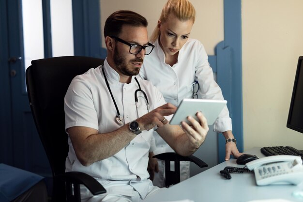 Médecin de sexe masculin et infirmière coopérant lors de l'examen des dossiers médicaux électroniques d'un patient dans le cabinet du médecin