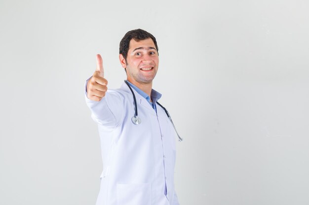 Médecin de sexe masculin montrant le pouce vers le haut en blouse blanche et regardant joyeux. vue de face.