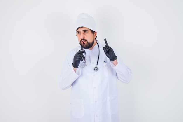 Médecin de sexe masculin mordant des lunettes tout en pointant vers le haut en uniforme blanc et en ayant l'air concentré. vue de face.