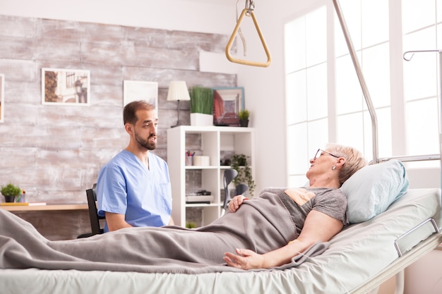 Médecin de sexe masculin parlant avec une femme âgée à la retraite dans une maison de soins infirmiers allongée dans son lit.
