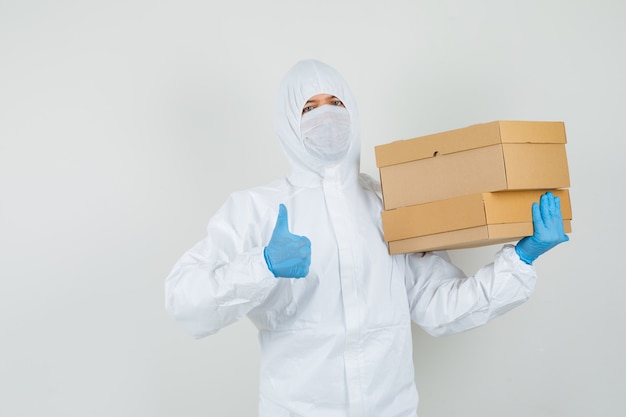 Médecin de sexe masculin tenant des boîtes en carton, montrant le pouce vers le haut en tenue de protection