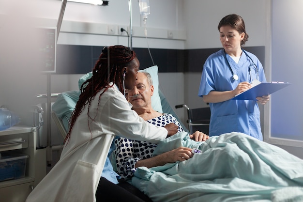 Médecin spécialiste africain utilisant un stéthoscope écoutant le cœur d'un homme âgé allongé dans son lit respirant avec ...