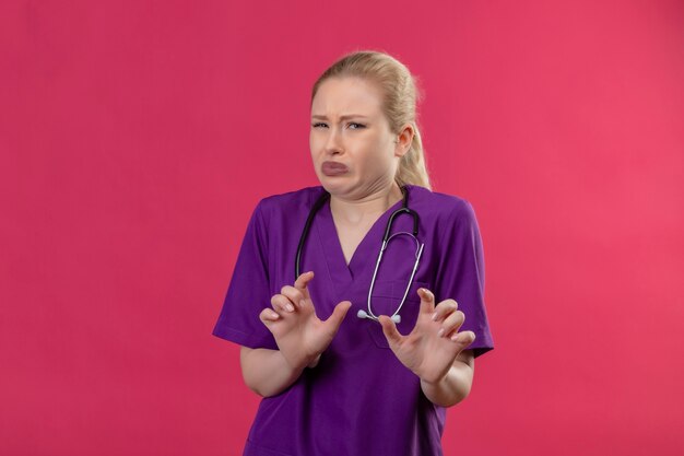Médecin squeamish jeune fille portant une robe médicale violette et stéthoscope sur fond rose isolé