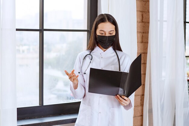 Médecin avec stéthoscope et masque noir debout à côté de la fenêtre et tenant un dossier d'histoire noire des patients, le vérifiant et a l'air terrifié.